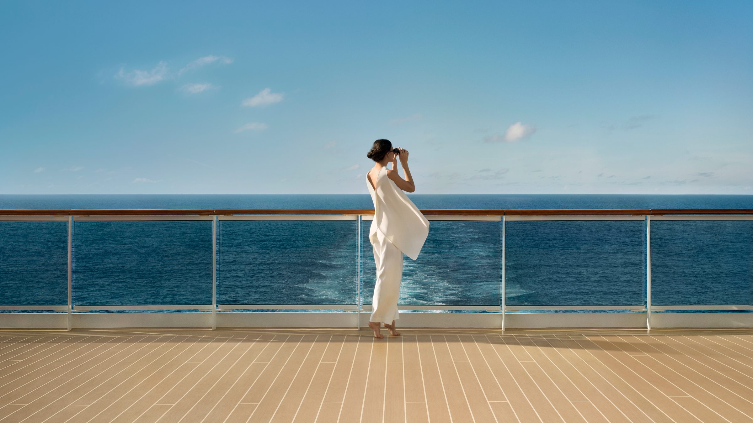 A traveler in white stands on the aft deck, looking through binoculars toward an open horizon.