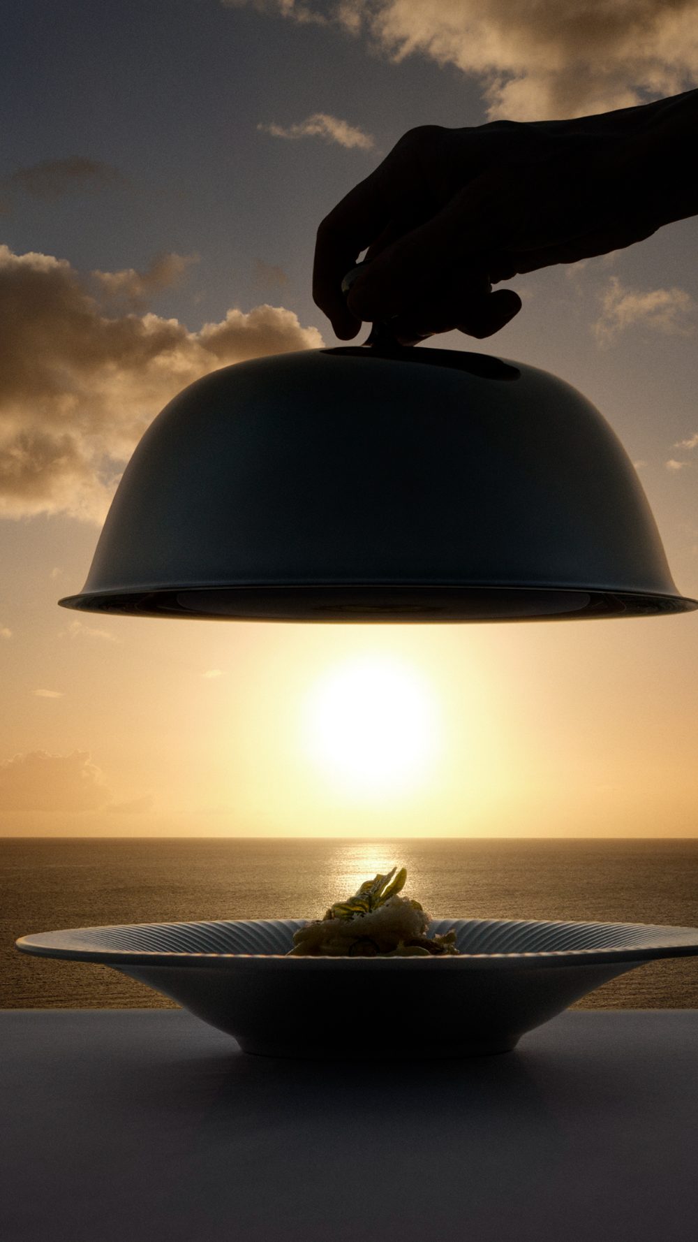 A server lifts a cloche to reveal a plated dish against the setting sun over the ocean.